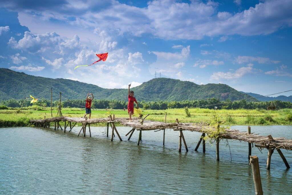 In the moment Two children playing with kites on a scenic wooden bridge over a river in a lush landscape.